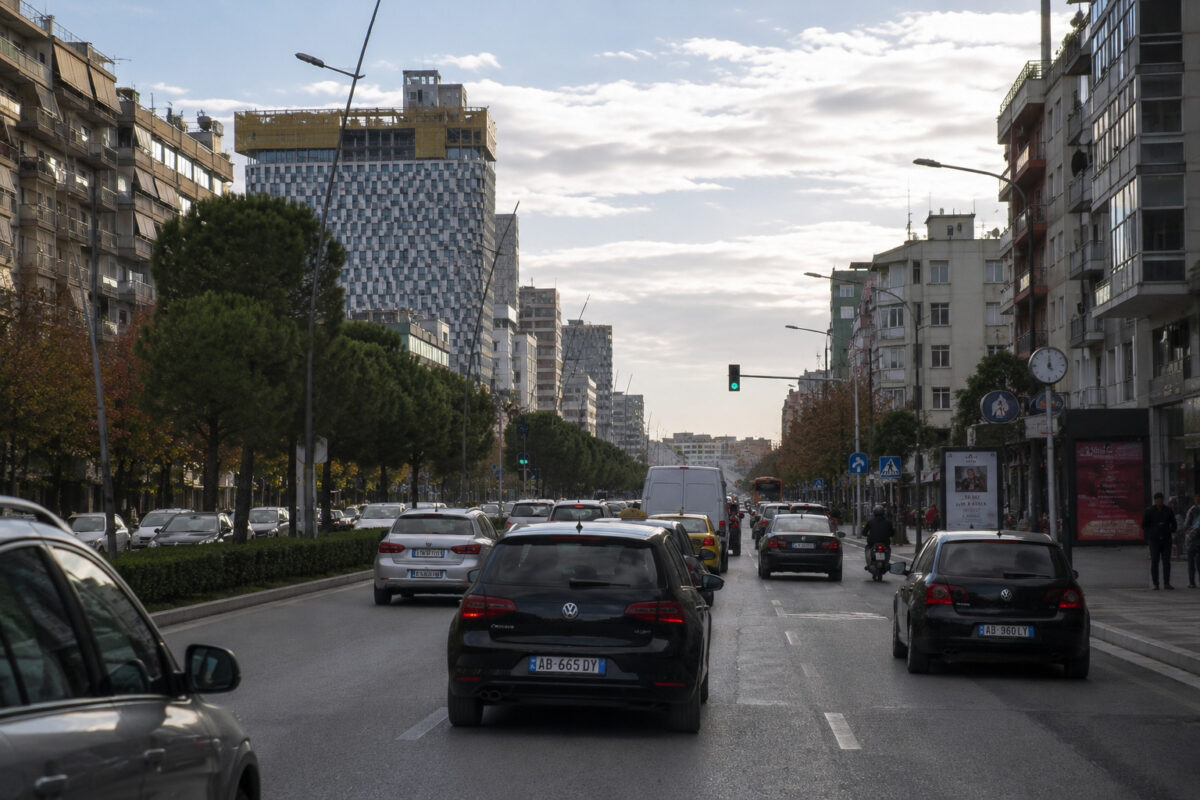 Calle urbana de Tirana con tráfico moderado, coches circulando, edificios residenciales y ambiente cotidiano de ciudad balcánica.