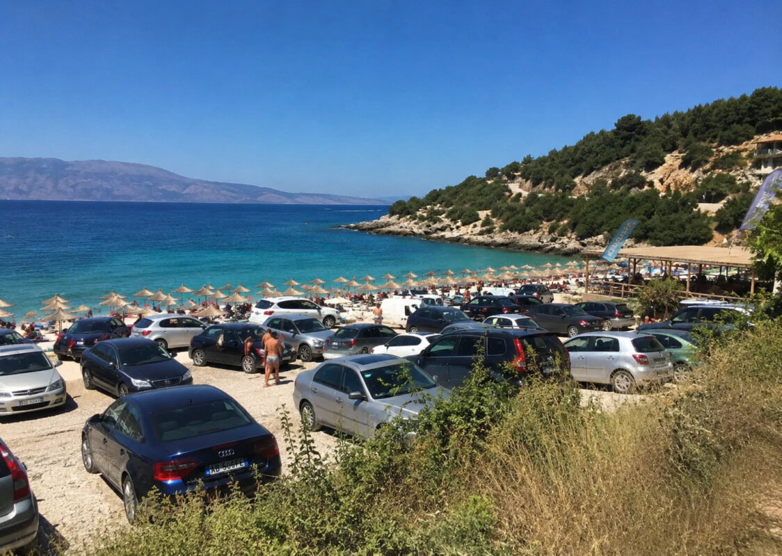 Parking lleno de coches junto a una playa de aguas turquesas en la Riviera albanesa, rodeada de montañas y vegetación mediterránea.